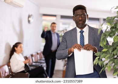 Angry, enraged boss yells at subordinate African-American male colleague in glasses and suit. In meeting room, African worker is upset because of boss abuse and tears up application document statement - Shutterstock ID 2310557389