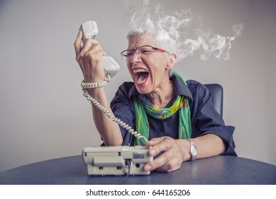 Angry, enraged senior woman yelling at a landline office phone, unhappy with customer service provided by the agent on the other side, giving off steam and smoke - Shutterstock ID 644165206