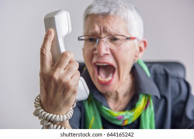 Angry, enraged senior woman yelling at a landline office phone, unhappy with customer service provided by the agent on the other side