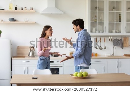 Angry excited caucasian millennial husband and wife quarreling, yelling at each other and gesturing in kitchen interior. Relationship problems, stress, negativity, divorce and scandal during covid-19