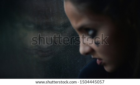 angry face of young little caucasian girl wearing dark youth subculture makeup watching at her sinister reflecton in window
