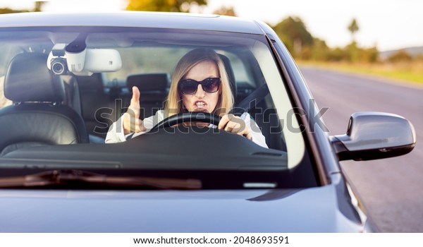 Angry female driver shouting and\
gesturing while driving modern car. Stressed business lady wearing\
sunglasses and formal clothes. Negative human\
emotions.