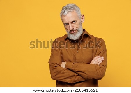 Angry frowning elderly gray-haired bearded man 40s years old wears brown shirt looking camera hold hands crossed isolated on plain yellow background studio portrait. People emotions lifestyle concept