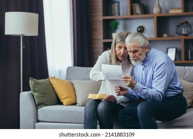 Angry Frustrated Document Senior Couple. Unhappy Old Family Getting Stressed. Serious Older Husband Showing Banking Paper Document Bills Taxes To Confused Elderly Wife In Home. Worried Looking Utility