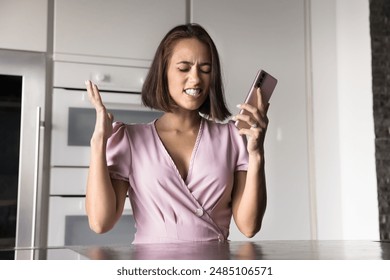 Angry frustrated young woman getting bad news, problems with mobile phone work, holding smartphone, making mad hands gesture, feeling shocked, annoyed, standing in home kitchen - Powered by Shutterstock