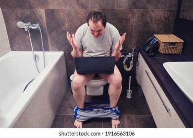 Angry Fun Man In Casual Clothes Works While Sitting In The Home Toilet. Problems With Telecommuting From Home During A Virus Pandemic