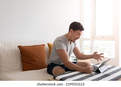 Angry And Furious Asian Trader Man Working On Tablet Or Laptop In His Apartment.