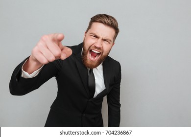 Angry furious businessman in suit shouting and pointing finger at camera isolated over gray background
