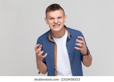 Angry furious teenager boy wearing blue shirt standing and screaming with anger, expressing aggression, swearing with someone, having problems. Indoor studio shot isolated on gray background. - Shutterstock ID 2310458553