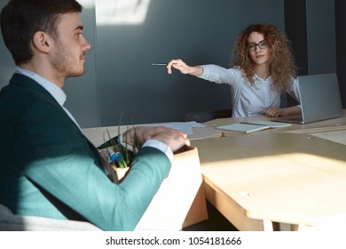 Angry Furious Young Woman CEO In Glasses And Shirt Sitting In Front Of Open Laptop At Her Workplace, Holding Pen And Pointing At Door While Firing Incompetent Man Worker For Bad Work Results