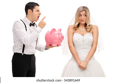 Angry groom holding a piggybank and scolding an embarrassed bride isolated on white background