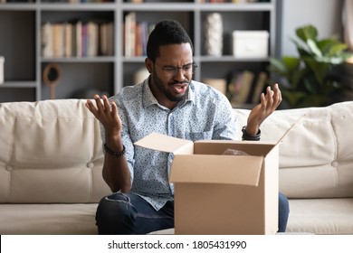 Angry irritated African American man in glasses dissatisfied by received parcel, unhappy customer unpacking cardboard box, wrong or broken fragile online store order, bad delivery service concept