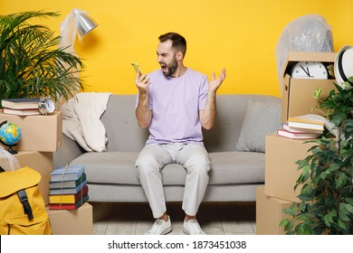 Angry irritated owner man talking on mobile phone screaming swearing sits in living room on sofa at home unpacking stuff rents flat isolated on yellow wall. Relocation moving in apartment concept - Shutterstock ID 1873451038