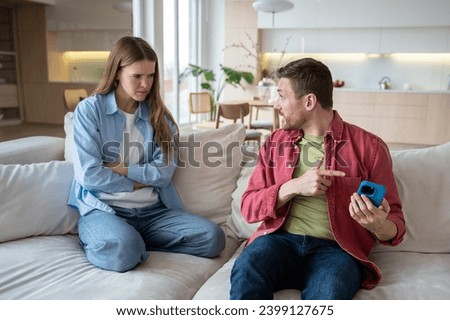 Angry irritated woman looking at man hiding smartphone display. Nervous wife listening explanations of sly justifying unfaithful husband who doesnt want to show mobile phone. Mistrust in family life