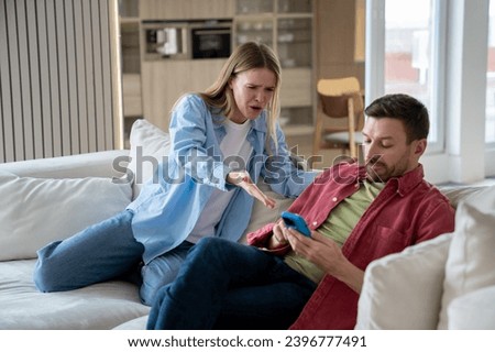 Angry irritated woman raising voice, shouting at demonstrative uncaring man sitting on sofa with mobile phone. Wife reproaches husbund in doing nothing, sitting and playing mobile games whole day