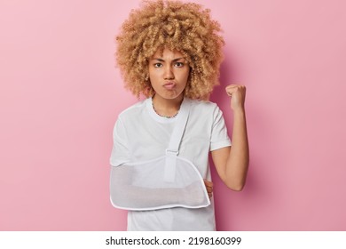 Angry irritated young woman with curly hair clenches fist feels annoyed wears sling in broken arm pouts lips isolated over pink background. Displeased female model has injury after accident.