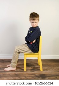 An Angry Little Boy Sits On A Yellow Time Out Chair On A Wooden Floor