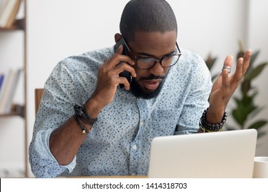 Angry mad african-american businessman talking on cellphone looking at laptop in office. Shocked manager received bad news discussing failing profit statistics with coworker, solving problem distantly