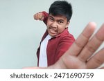 The angry and mad face of Asian man in red shirt and want to fight on isolated gray background.