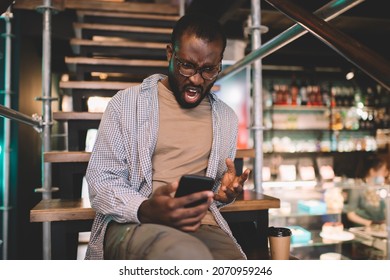 Angry Male Blogger In Optical Eyewear Screaming While Reading Email Message During Mobile Texting, Unhappy Dark Skinned Man In Spectacles Solving Problems With Internet Connection On Cellphone