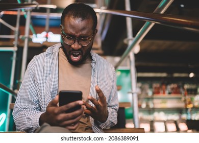 Angry Male Blogger In Optical Eyewear Screaming While Reading Email Message During Mobile Texting, Unhappy Dark Skinned Man Solving Problems With Internet Connection On Cellphone Feeling Stress