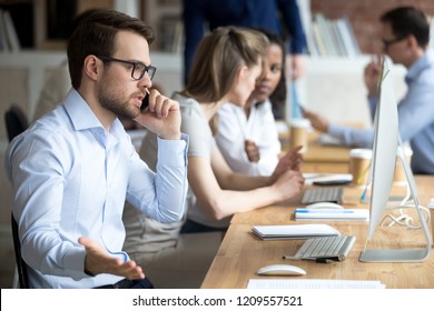 Angry male employee confused working at computer, solve business problems on phone, mad worker talk on cell arguing with client or customer, man busy at pc having serious conversation on cellphone