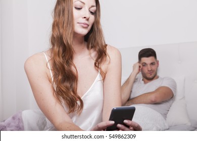 angry man in bed watches woman reading on smart phone