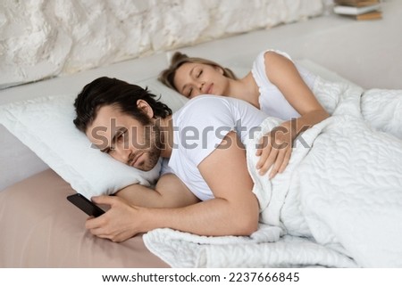 Angry man checking his sleeping girlfriend's smartphone, lying in bed at home. Husband feeling jealous of her wife's infidelity, looking through her phone messages