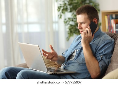 Angry man having problems on line with a laptop talking on the mobile phone with support service sitting on a couch in the living room at home