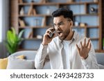 Angry man shouting on phone at home expressing frustration and stress. Young adult with glasses having heated conversation displaying intense emotions in modern living room setting.