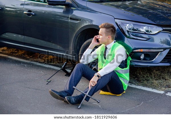 Angry\
man sitting down next to his car, calling for assistance, man in a\
green safety vest talking on cell phone. has problems with the\
wheel of his car. man sitting down next to his\
car,