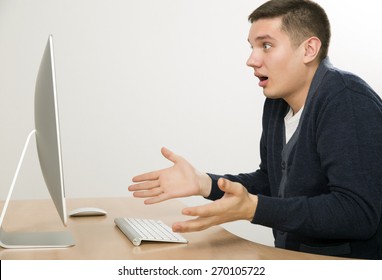Angry man yelling on computer screen. Young man expresses anger fury sitting in front of large computer screen. Smart casual dress beige office desk, wireless keyboard and mouse, light grey background