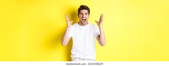 Angry man yelling and shaking hands, grimacing with hatred, standing hateful against yellow background. Copy space - Shutterstock ID 2321458229