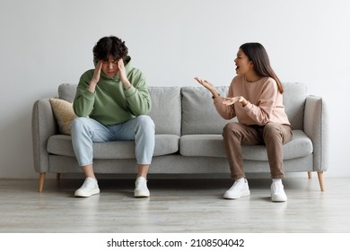 Angry Millennial Asian Wife Yelling At Her Husband, Man Suffering From Stress, Sitting On Sofa In Living Room, Free Space. Relationship Problems, Crisis, Divorce And Breakup Concept