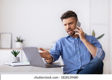 Angry millennial businessman talking on mobile phone and looking at laptop screen working in modern office, having problems with project, annoyed boss speaking with employee by phone, copy space - Shutterstock ID 1828139480