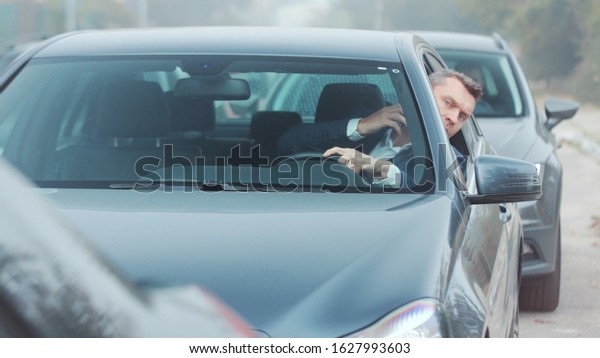 Angry nervous bearded man in luxury suit stands
in traffic jam, picks up his phone, answers call, explaining
situation, actively gesticulating. Being in a hurry, being late,
stressful situation
