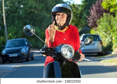 Angry Old Lady On A Scooter Making A Rude Gesture. Senior Woman Showing The Finger While Sitting On A Scooter Bike In A Sign Of Road Rage