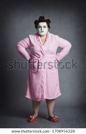 angry overweight woman with green beauty mask and hair curlers wearing pink bathrobe and slippers on gray studio background