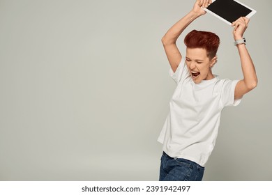 angry queer person in white t-shirt screaming and breaking digital tablet with blank screen on grey - Powered by Shutterstock