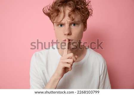Angry rigorous ginger curly haired man uses body language frowns brows displeased look shows please be quiet shush gesture presses finger to his lips asks not to tell the secret on pink backdrop.