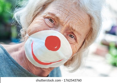 Angry Sad Grandmother Elderly Woman In A Respiratory Mask With A Painted Funny Clown Mask. Tired Of Wearing A Mask. Old Lady Makes Fun Of Wearing A Mask, Covid-19. Concept Of Deception Coronavirus