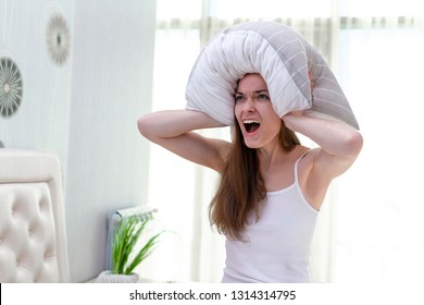 Angry screaming woman suffering and disturbed by noisy neighbors and covering her ears with pillow while trying to falling sleep in bed at home in early morning. 