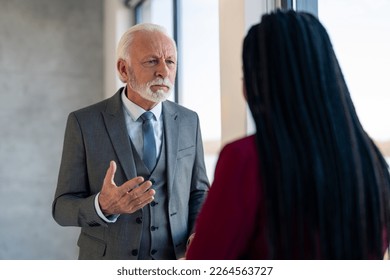 Angry senior executive manager in suit blaming and arguing with female coworker after misunderstanding while standing in office. Upset senior businessman looking angry at black haired businesswoman.