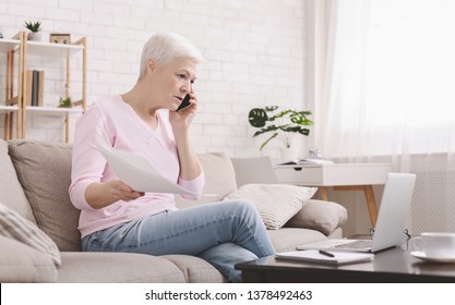 Angry senior woman talking on phone and working at home, looking at laptop and waving paper correspondence, sitting at home, free space