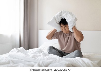 Angry sleepless nervous middle aged asian man with closed eyes sitting in bed and covering ears, head with pillow, hearing and suffering from too loud sound, tired of noisy neighbors, copy space