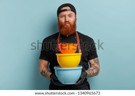 Angry strict male chef holds kitchenware, raises eyebrows, has thick ginger beard, wears cap and apron, buys new containers for kitchen, models over blue background. Cook with colorful dish.