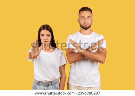 Angry strict millennial european man and lady in white t-shirts make stop gesture with hand, crossed arms, isolated on yellow studio background. End, fight with harassment, rejection