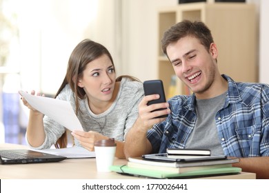 Angry student girl complaining showing homework to slacker classmate boy checking smartphone sitting on a desk at home