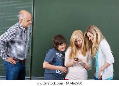 Angry Teacher Looking At Students Using Mobilephone Against Chalkboard In Classroom