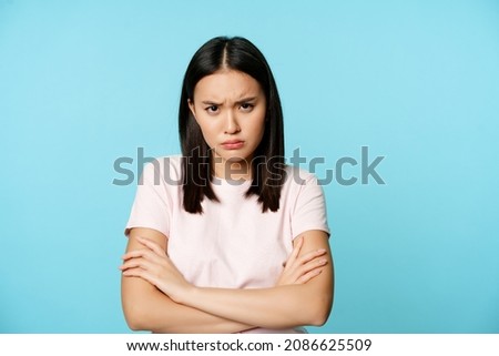 Angry teen asian girl standing in offended pose, frowning and sulking, feeling defensive, standing disappointed over blue background